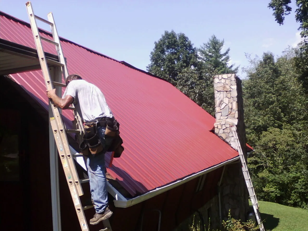 Roofer on a ladder beside a red metal roof and stone chimney — Soffit & Fascia Repair by Roofing Brothers in Gibson Island